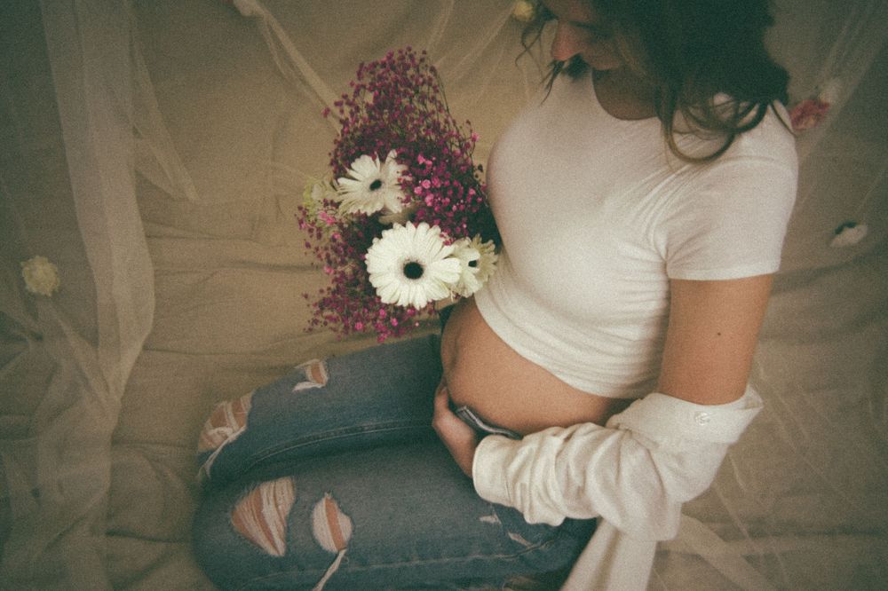 Pregnant woman in white top and ripped jeans sitting and holding white gerbera daisies against her baby bump.