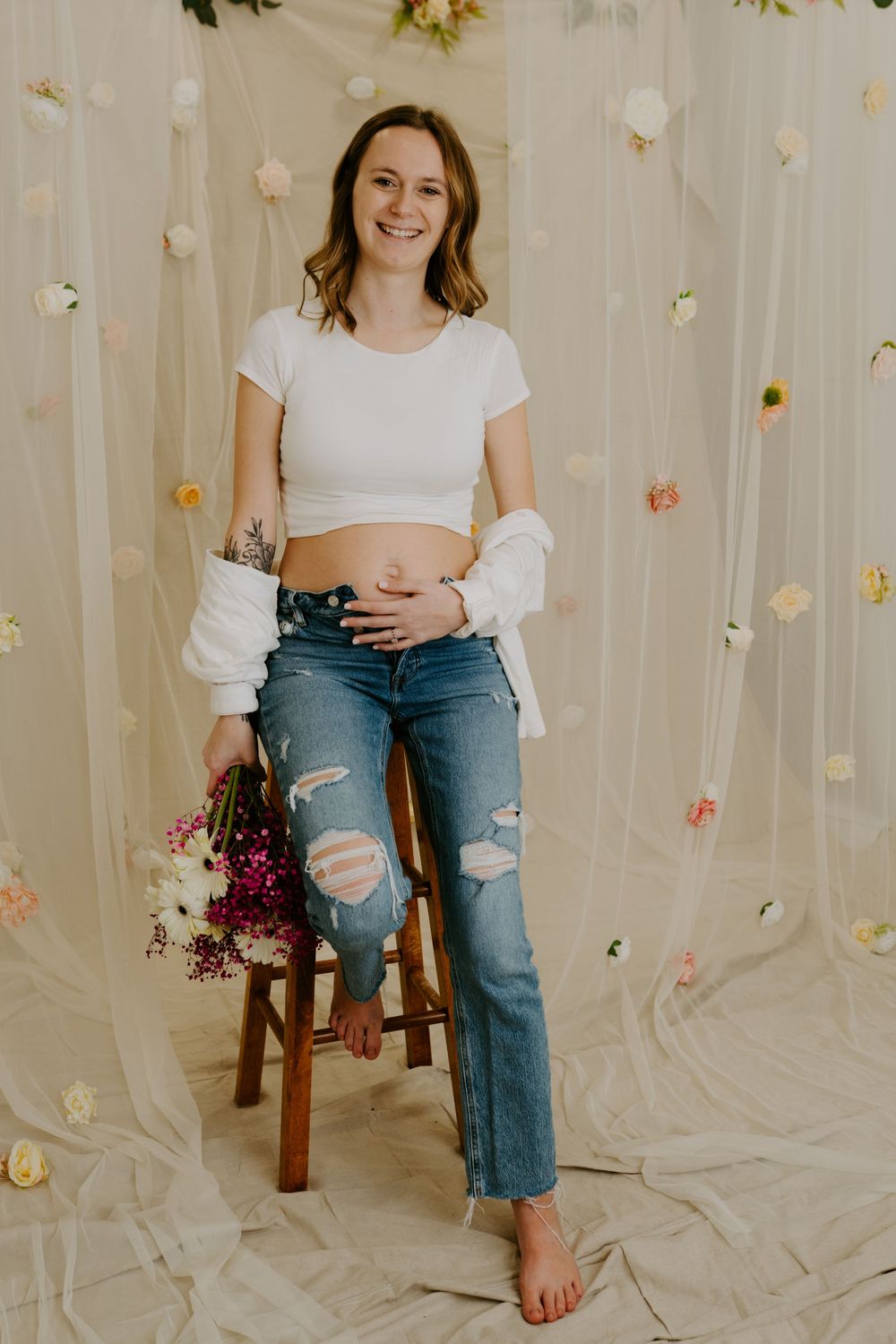 Smiling pregnant woman in white crop top and ripped jeans poses barefoot in front of floral backdrop.