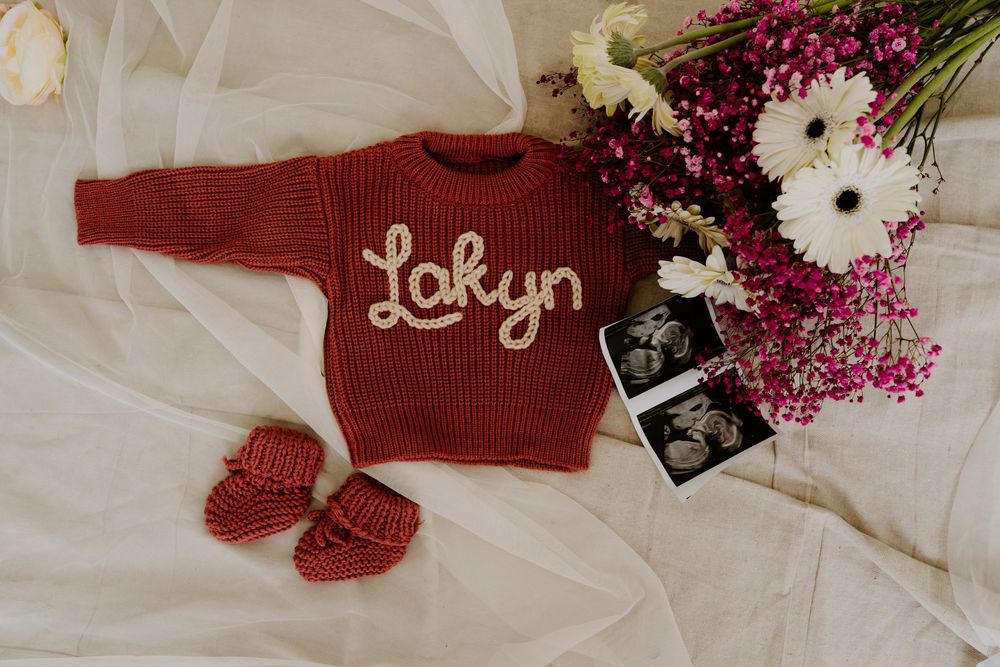 Red knit baby sweater personalized with name Lakyn, matching booties, ultrasound photos, and flowers on white fabric.