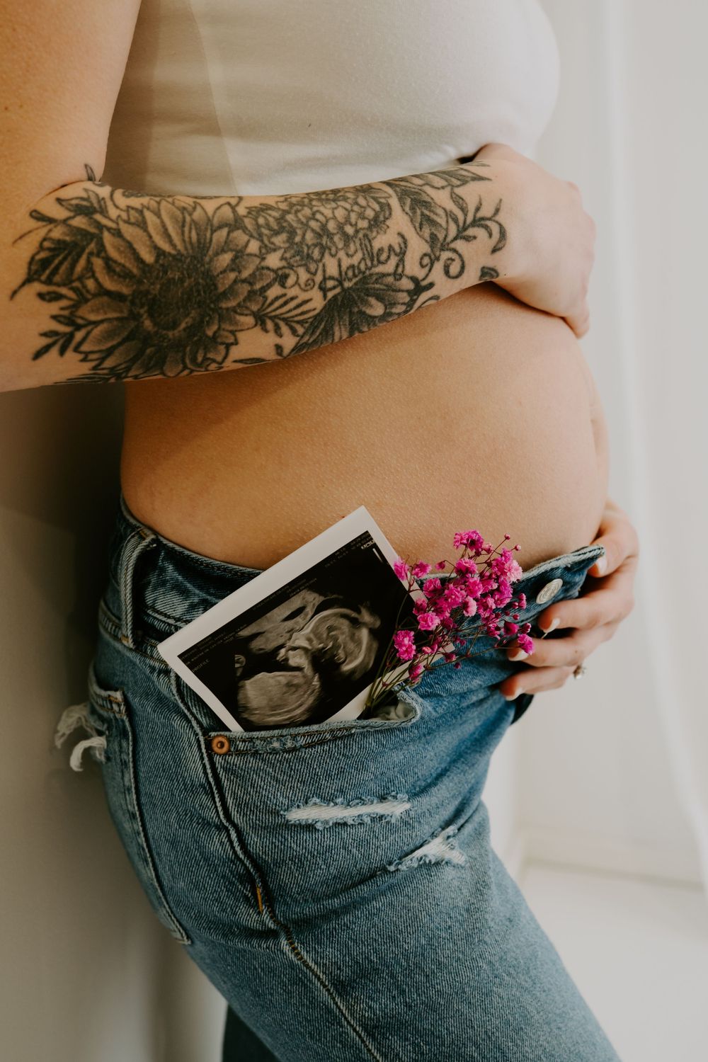 Pregnant woman with floral arm tattoo in jeans holds ultrasound photo and pink flowers near baby bump.
