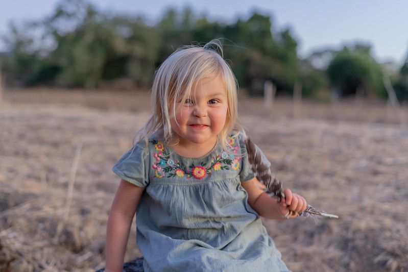 A young child wearing a blue dress smiles while sitting in a plowed field at sunset.