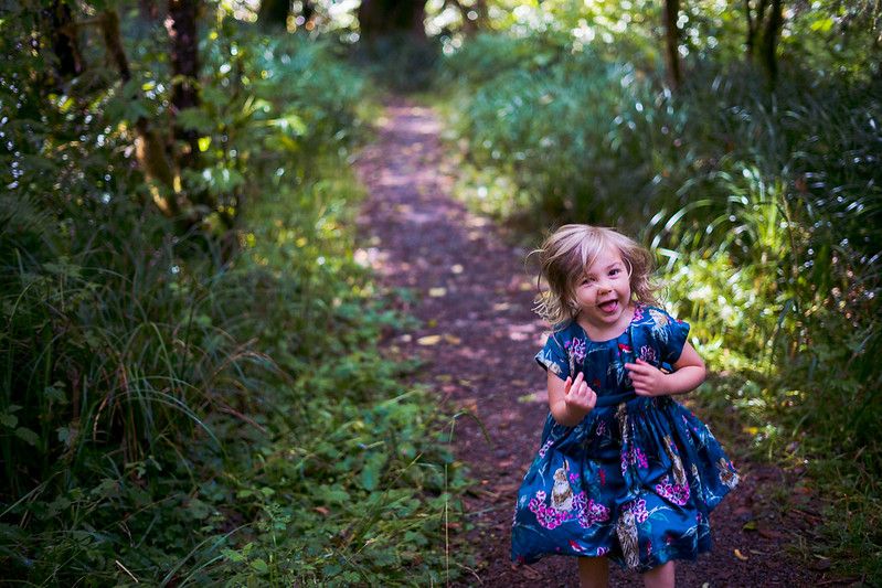 A small figure in a blue floral dress walks along a winding dirt path through lush green foliage.