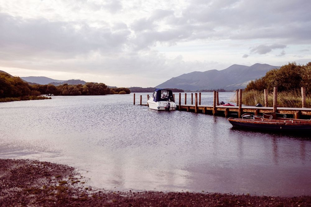 A serene lake with a wooden dock, moored boats, and misty mountains under a cloudy sky.