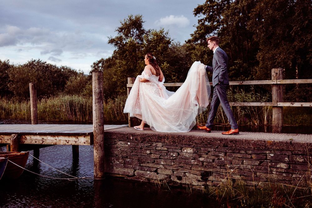Bride and groom walking on a stone bridge over water, surrounded by lush greenery on their wedding day.