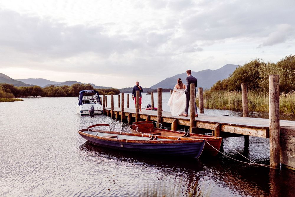 Wedding couple on a wooden dock by a serene lake with mountains, a rowboat nearby under cloudy skies.