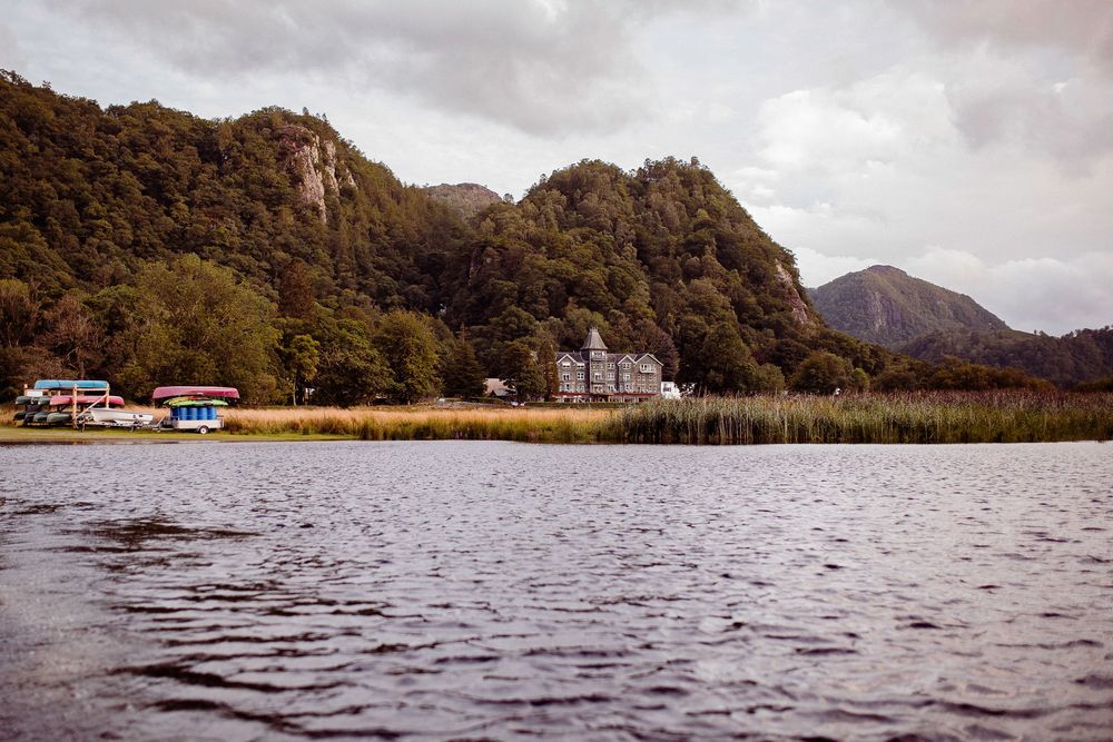 Scenic lake view with lush green mountains, reed beds, and a historic building nestled along the tranquil shoreline.