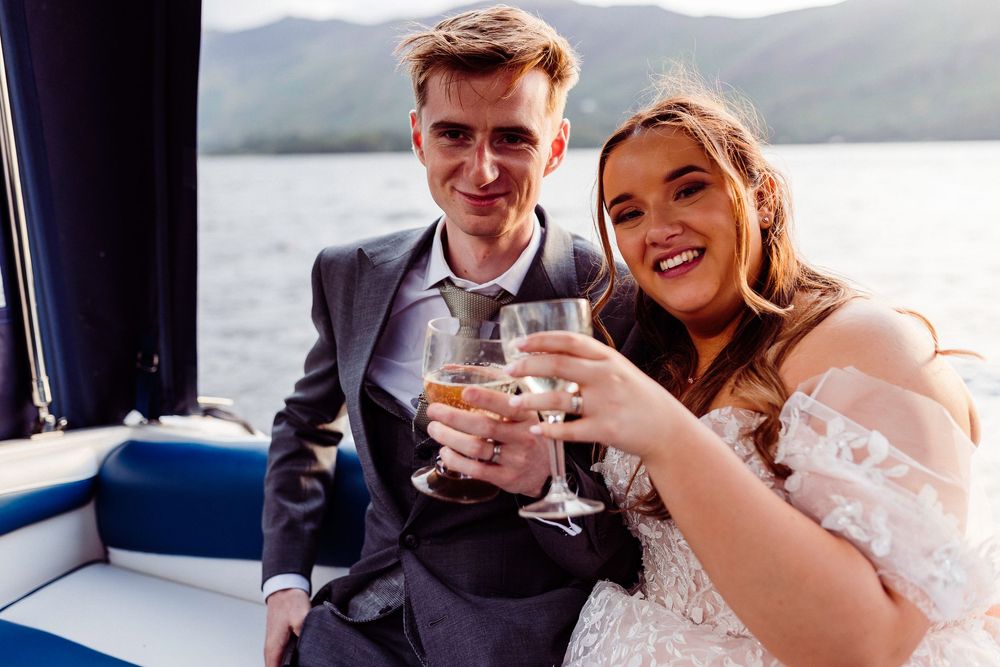 Newlywed couple toasting with champagne glasses on a boat, bride in lace gown and groom in gray suit.