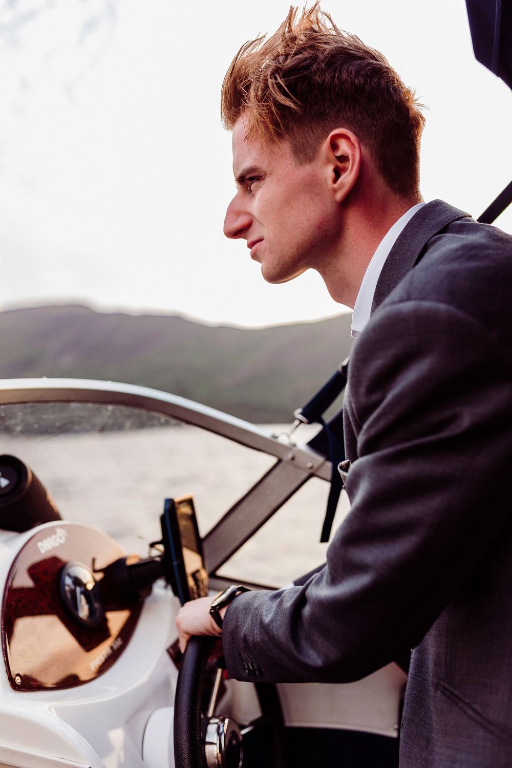 Young businessman in gray suit steering a luxury motorboat on a lake with mountains in the background.