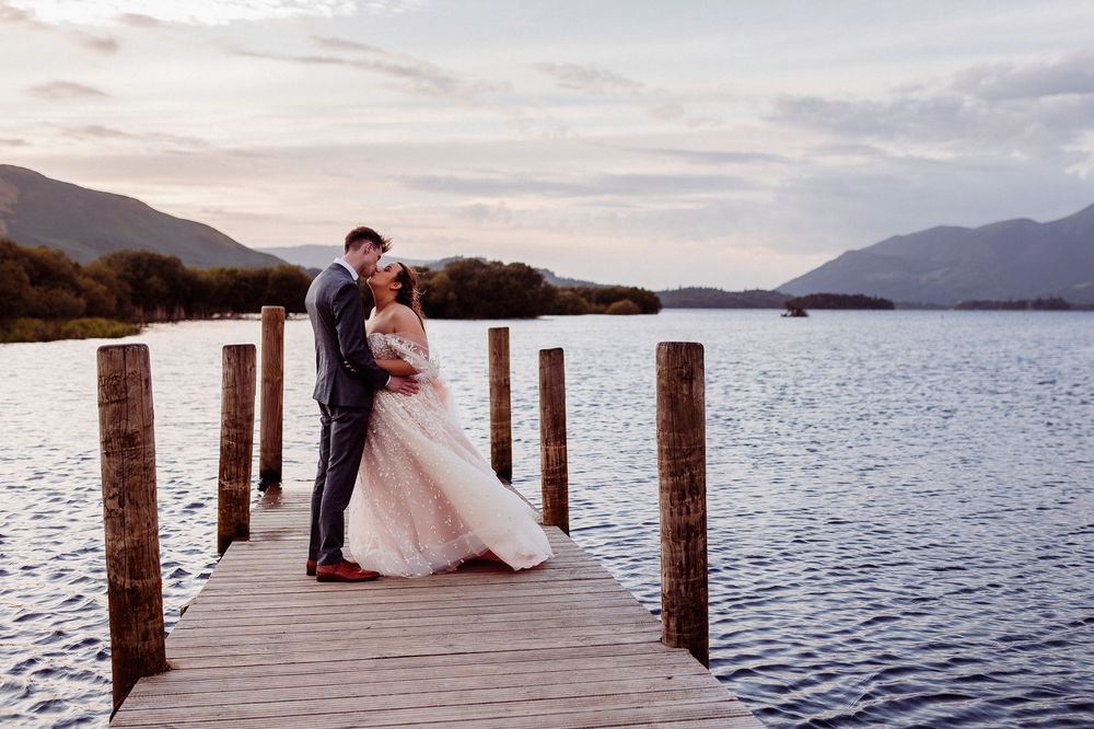 Bride and groom sharing a romantic kiss on a lakeside dock with mountain backdrop at sunset.