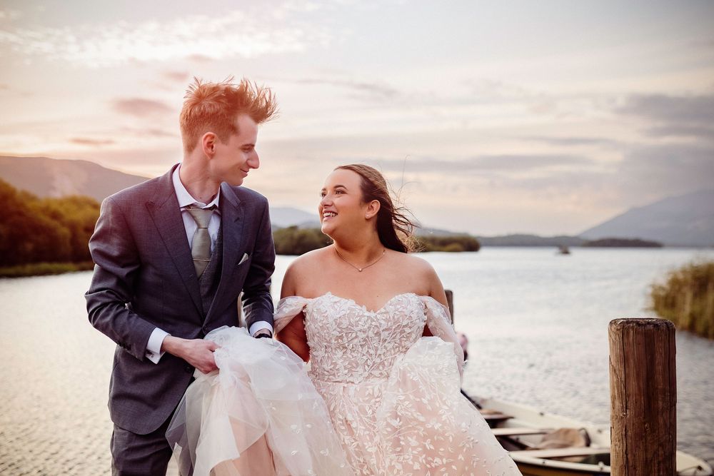 Bride and groom share romantic moment on lakeside dock at sunset in elegant wedding attire.