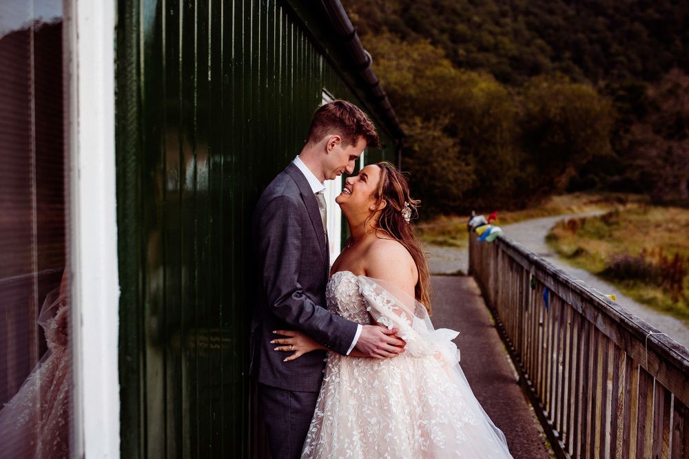 Bride and groom share a romantic kiss leaning against a green wall on a wooden deck in a scenic outdoor setting.