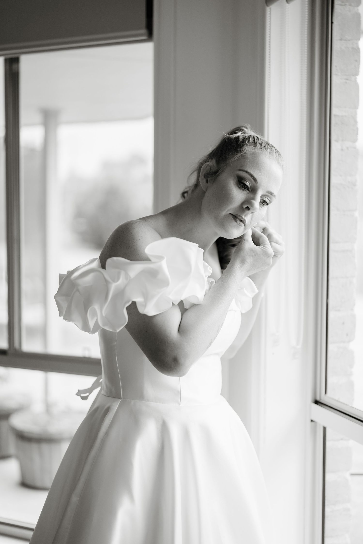 Black and white photo of a bride in an off-shoulder ruffled wedding gown posing elegantly by a sunlit window.