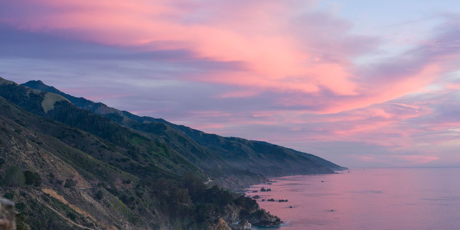 Vibrant pink sunset clouds illuminating Big Sur coastline with rugged mountains descending into calm purple ocean.
