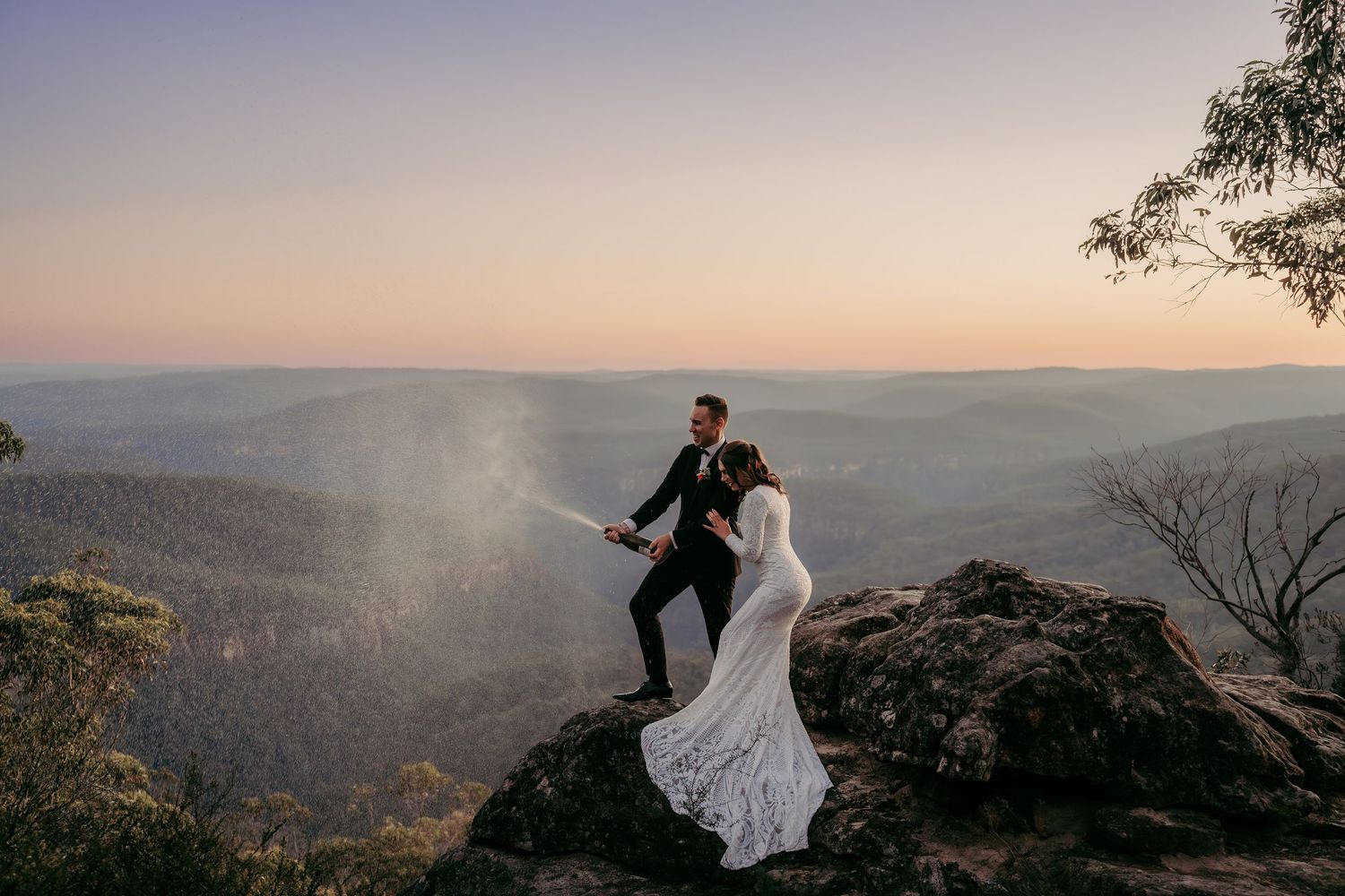 Bride and groom celebrate on rocky mountain peak at sunset with stunning valley views in Australian wilderness.