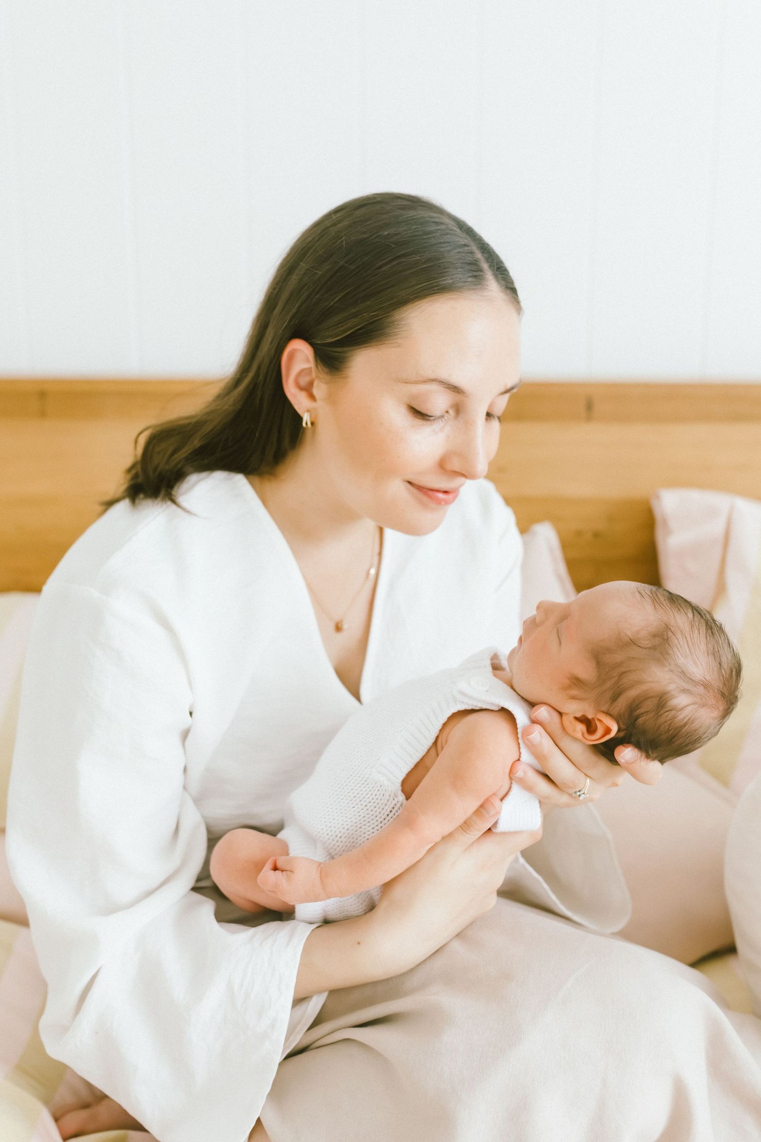 A smiling mother in a white robe tenderly holds her newborn baby while sitting on a bed.