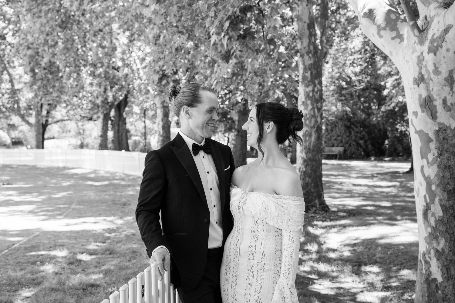 Black and white photo of a couple in wedding attire sharing a romantic moment among trees in a sunlit park.