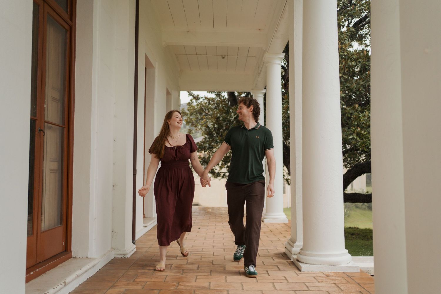 Couple holding hands and walking along a white columned veranda on a brick pathway. Engagement photoshoot in sydney watsons bay