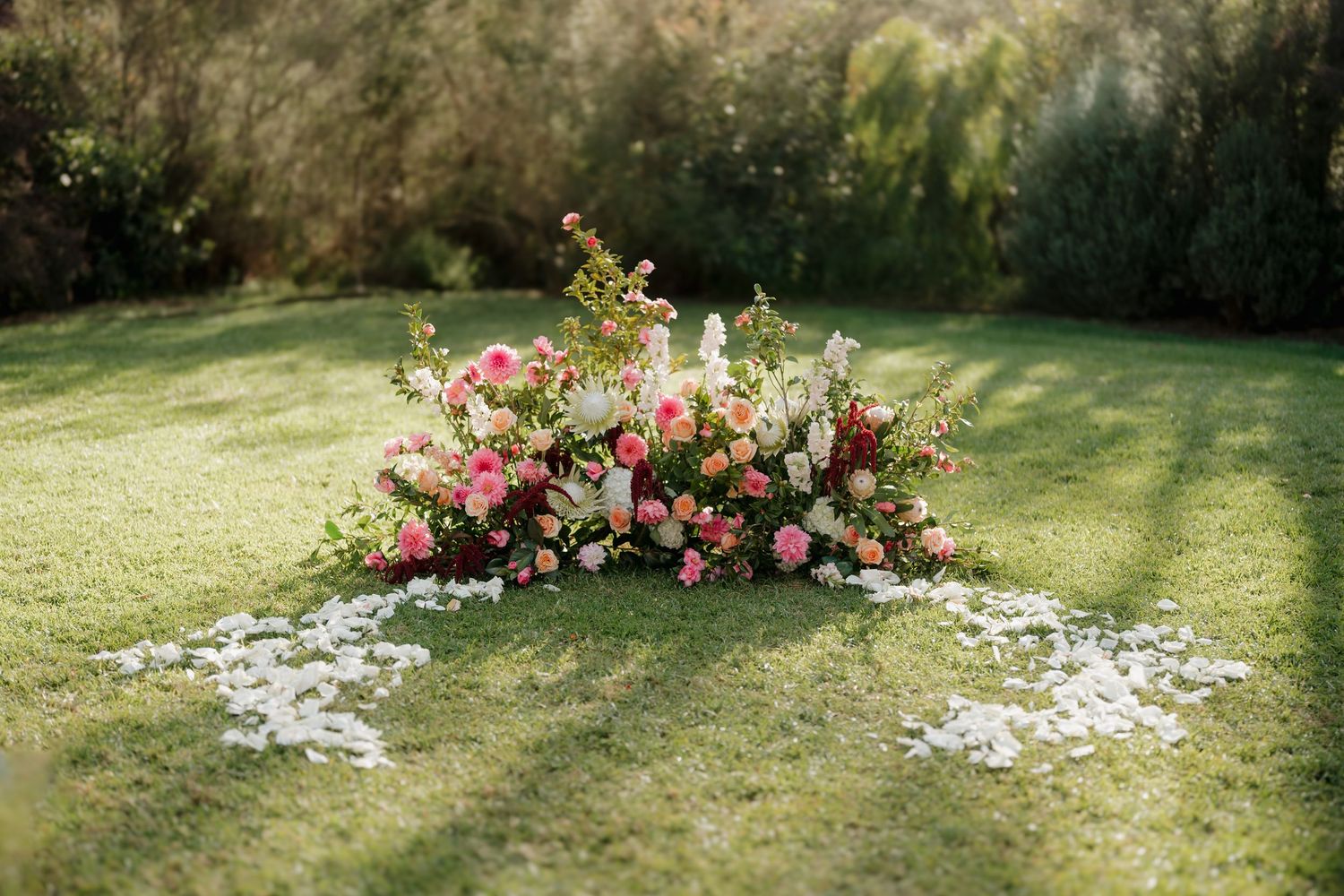 Lush floral arrangement of pink, coral, and white blooms on green grass with scattered petals at an outdoor wedding ceremony.