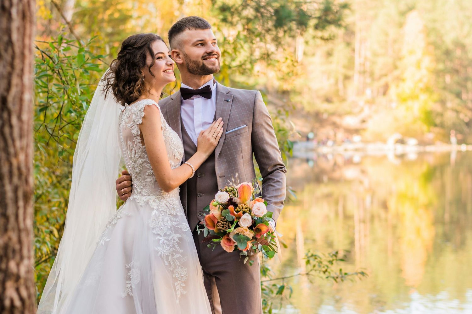 Les mariés posent de façon romantique au bord d'un lac forestier baigné de soleil, elle en robe de dentelle et bouquet, lui en costume gris