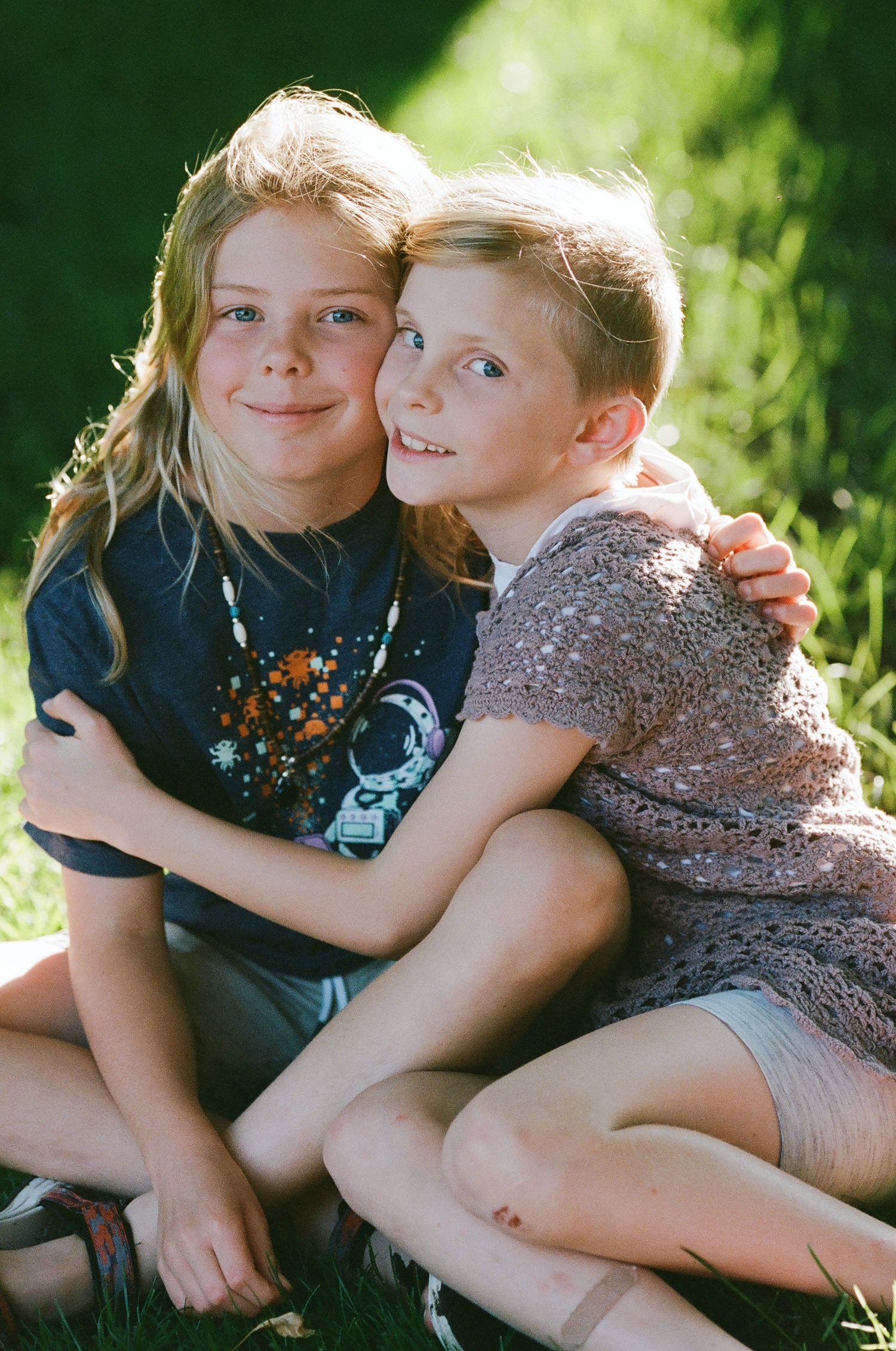 Two smiling children posing together on green grass in a garden, wearing casual summer outfits.