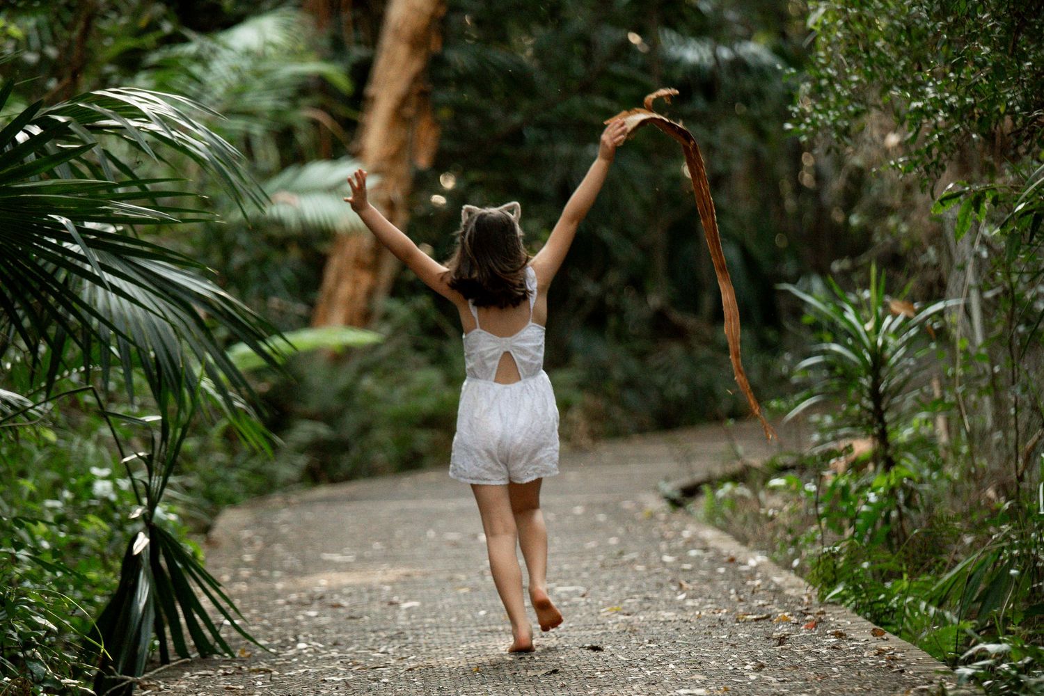 A young girl runs joyfully down a lush tropical garden path, arms raised holding a ribbon, wearing a white romper.