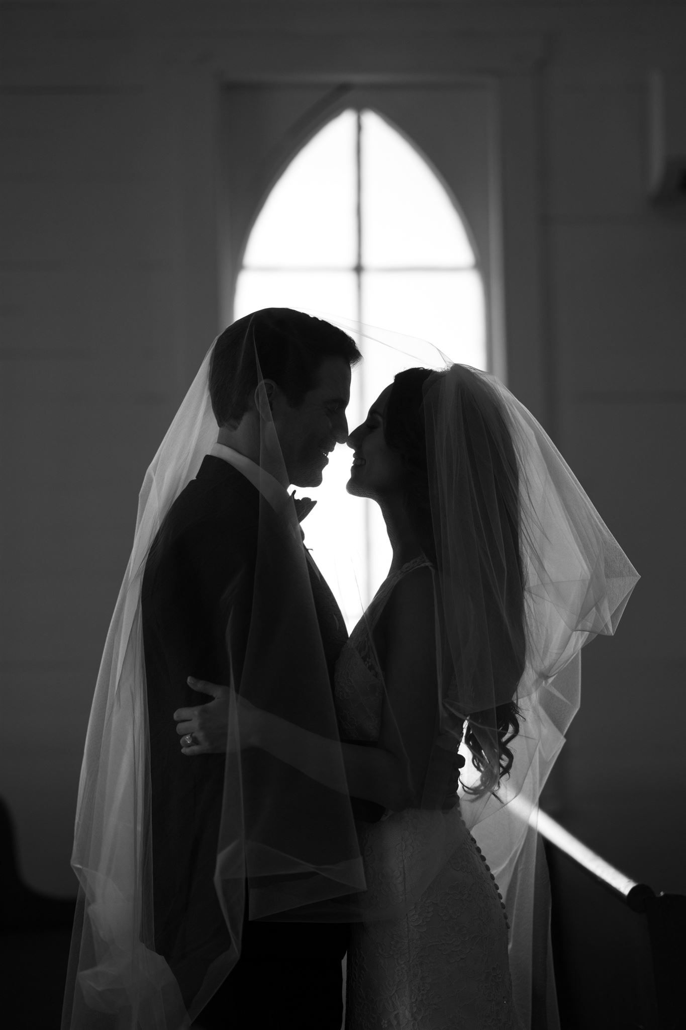 Black and white silhouette of bride and groom sharing romantic kiss under veil in historic Tomball chapel.