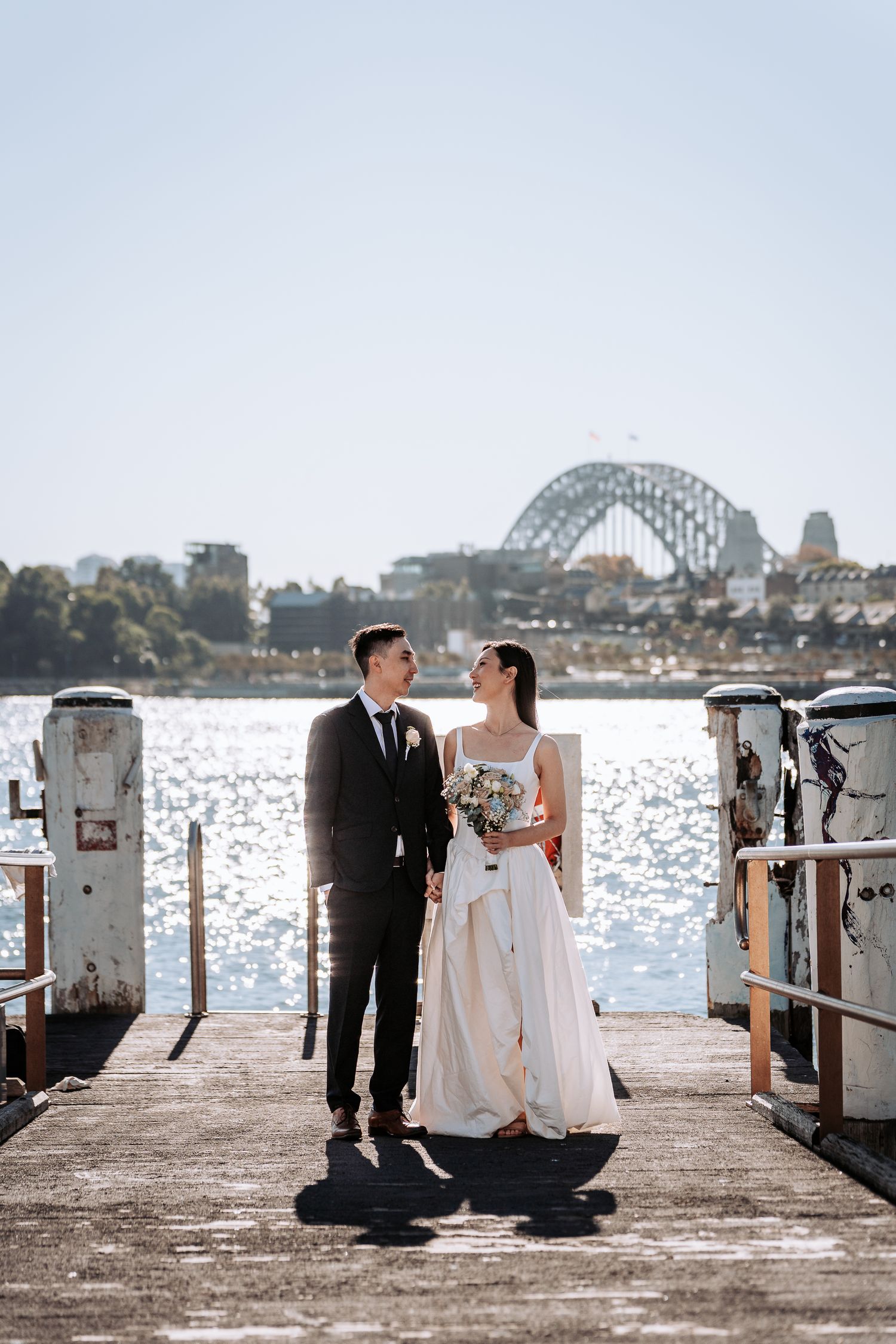 Bride and groom share a romantic moment on a Sydney harbour pier with the iconic Harbour Bridge in the background.