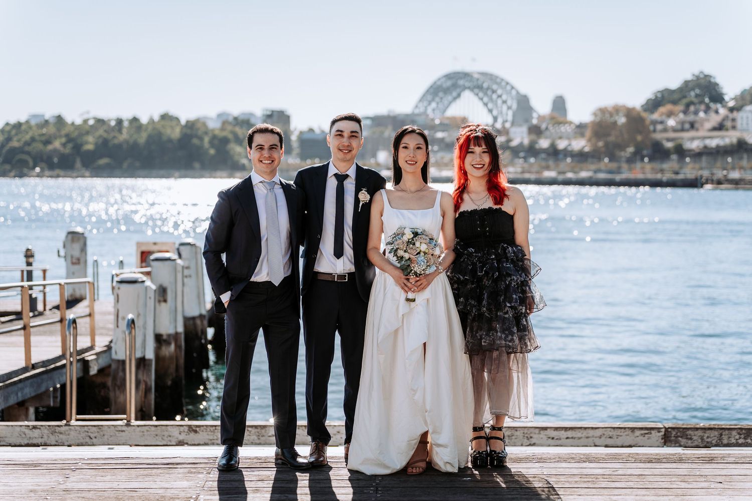 Wedding party of four posing on a waterfront dock with Sydney Harbour Bridge in the background on a sunny day.
