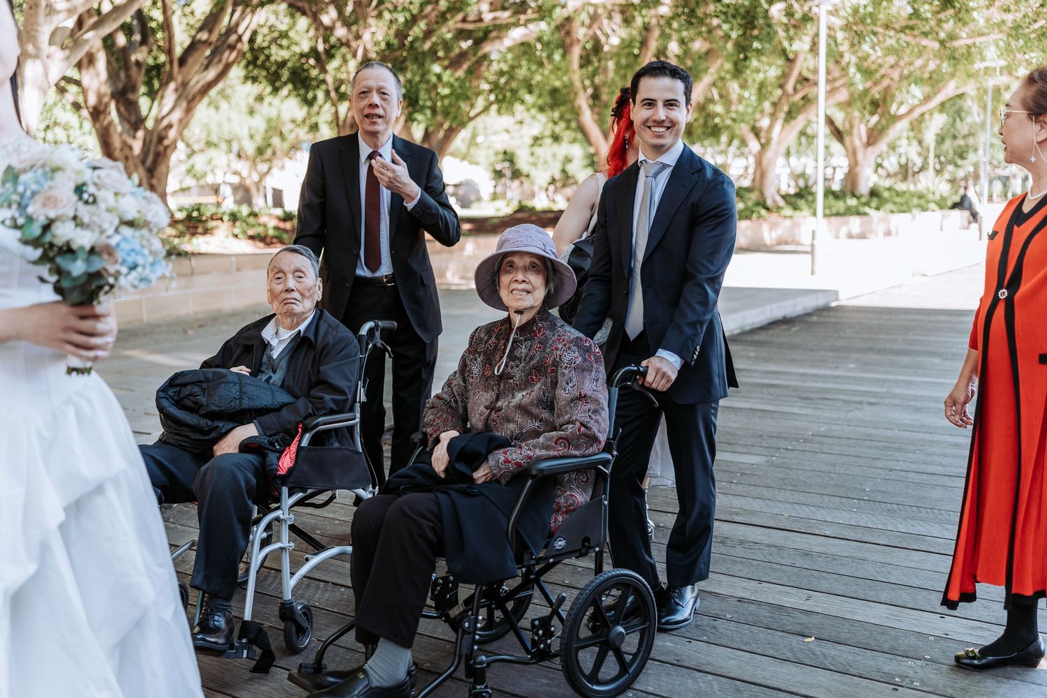 Wedding guests including two elderly people in wheelchairs smile outdoors at a sunny park ceremony.