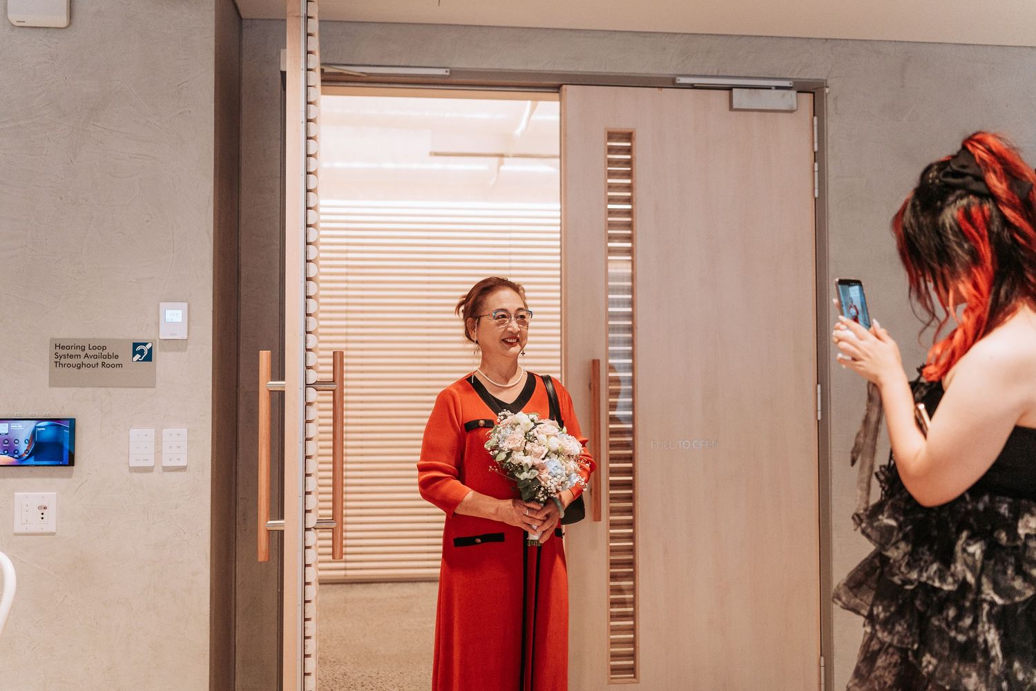 Woman in red dress holding white bouquet smiles as another woman photographs her near glass doors.