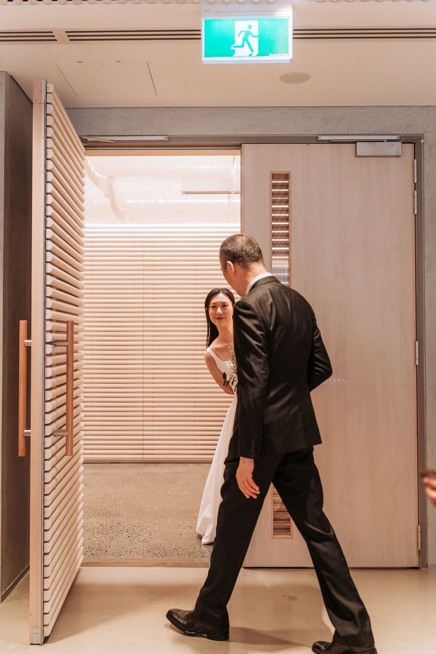 Groom walks toward bride in a modern venue corridor as she looks back with a smile.