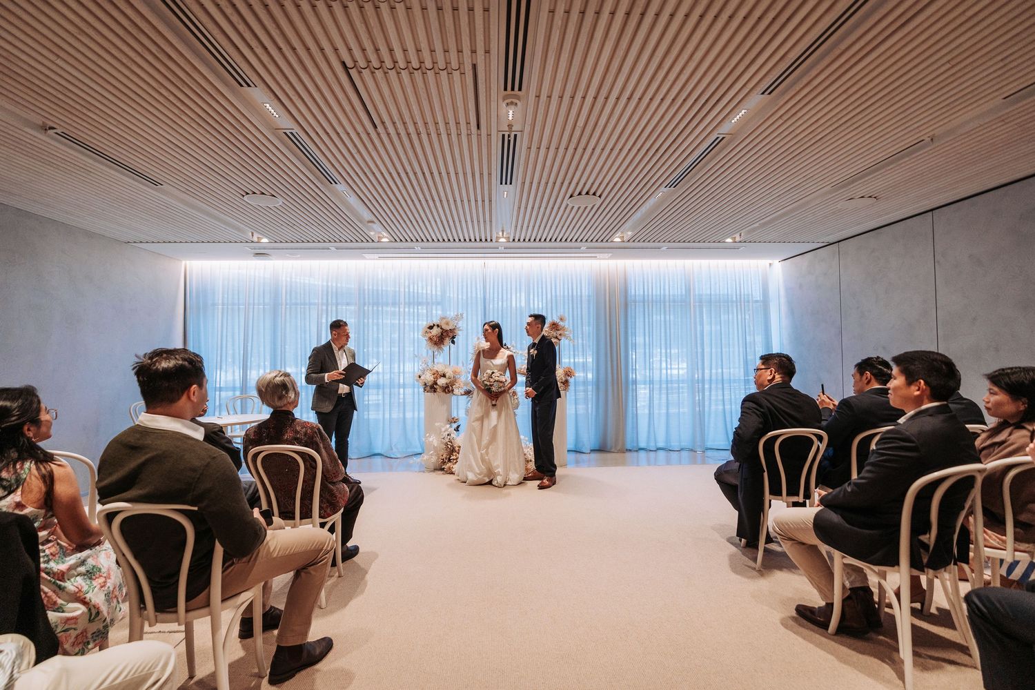 Couple exchanges vows during an elegant indoor wedding ceremony with seated guests in white chairs.