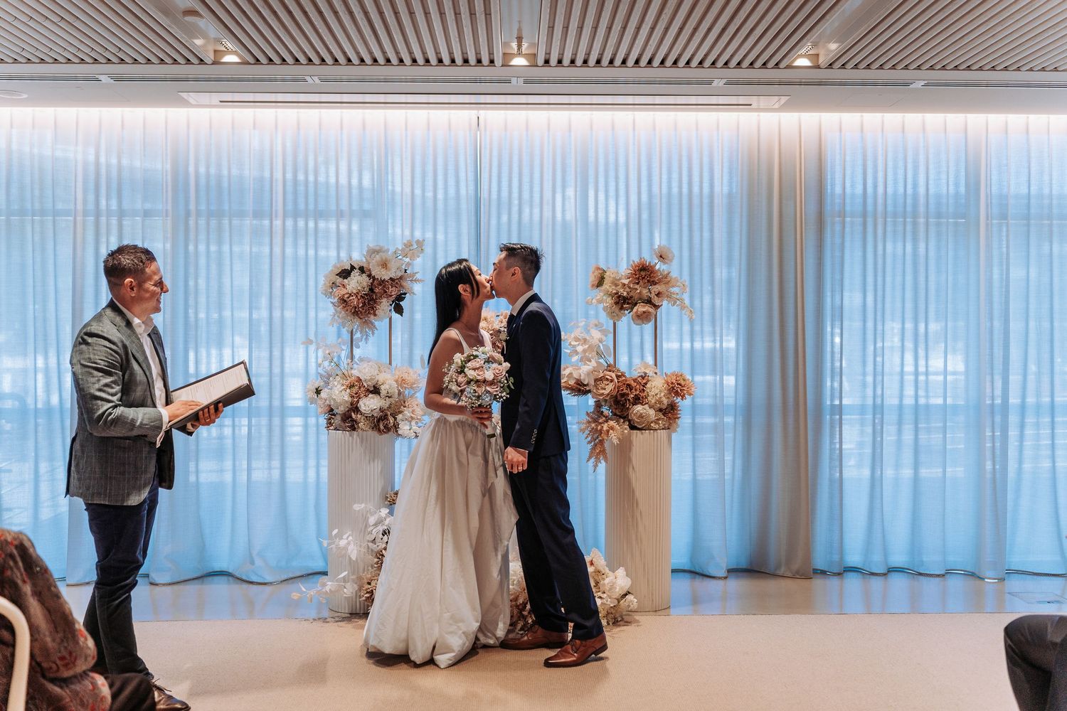 Couple exchanging vows at elegant indoor wedding ceremony with floral arrangements and blue curtains.