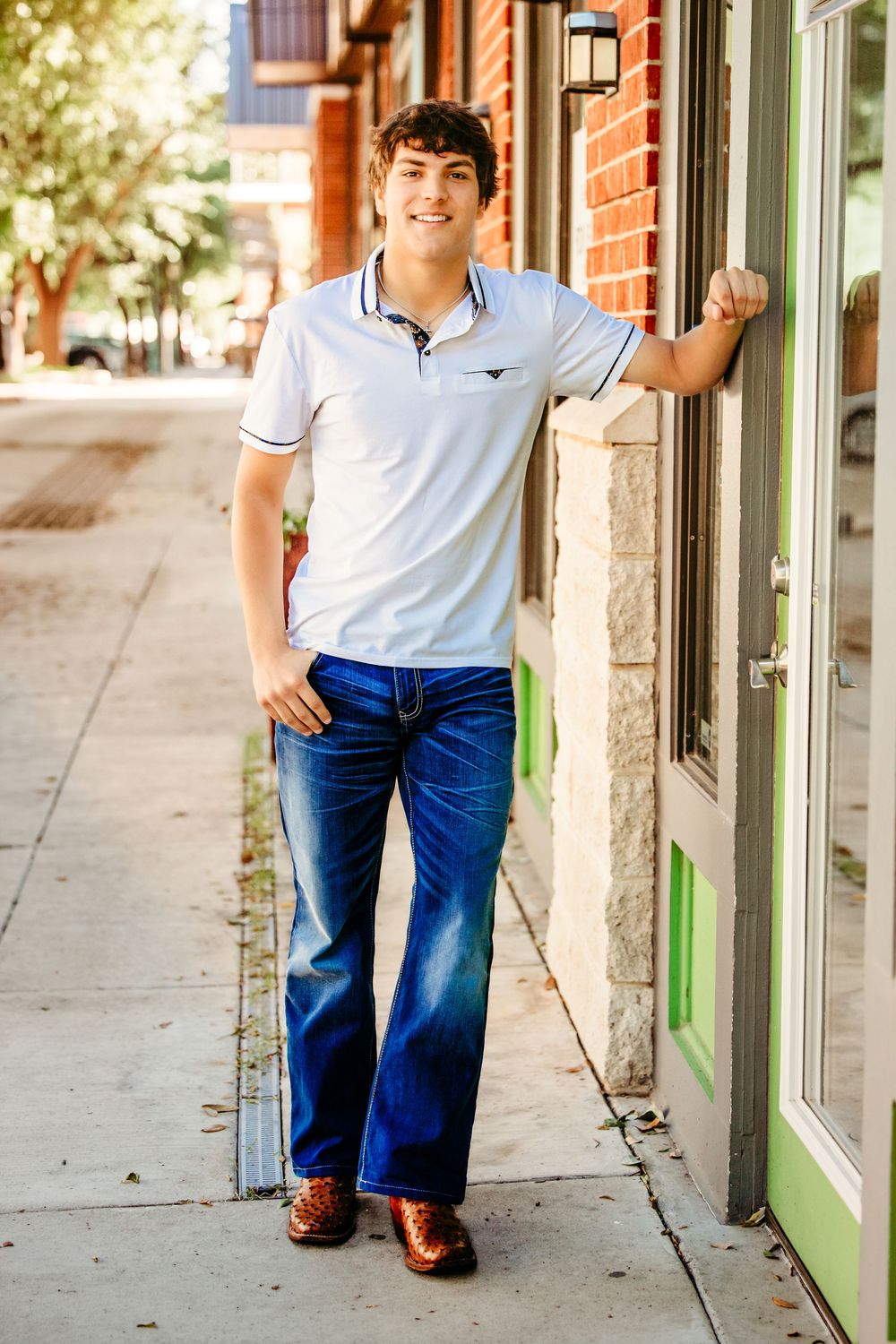 Young man in white polo shirt and jeans smiling while leaning against a storefront on a sunny sidewalk in Keller