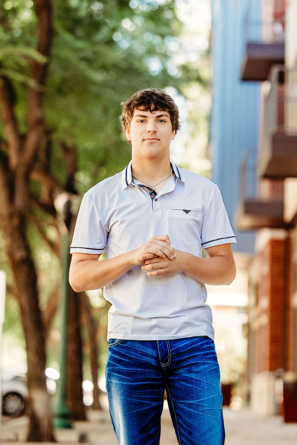 Young man in white polo shirt and blue jeans standing on urban sidewalk with trees in background in Keller