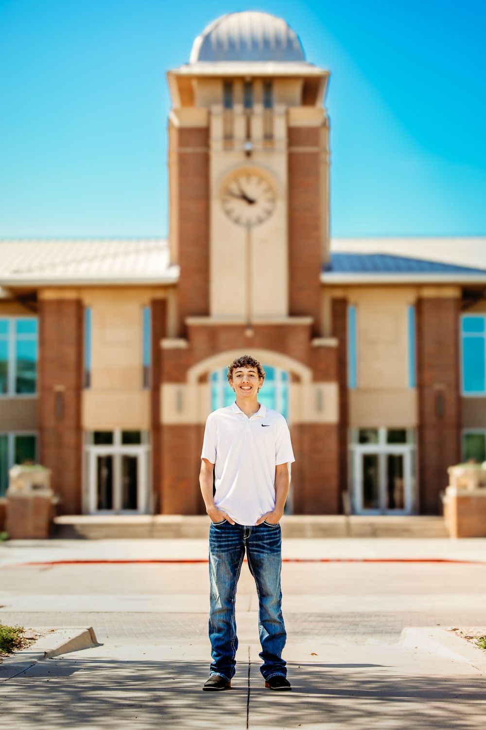 Young man in white Nike polo and jeans smiling in front of a brick school building with clock tower in Keller