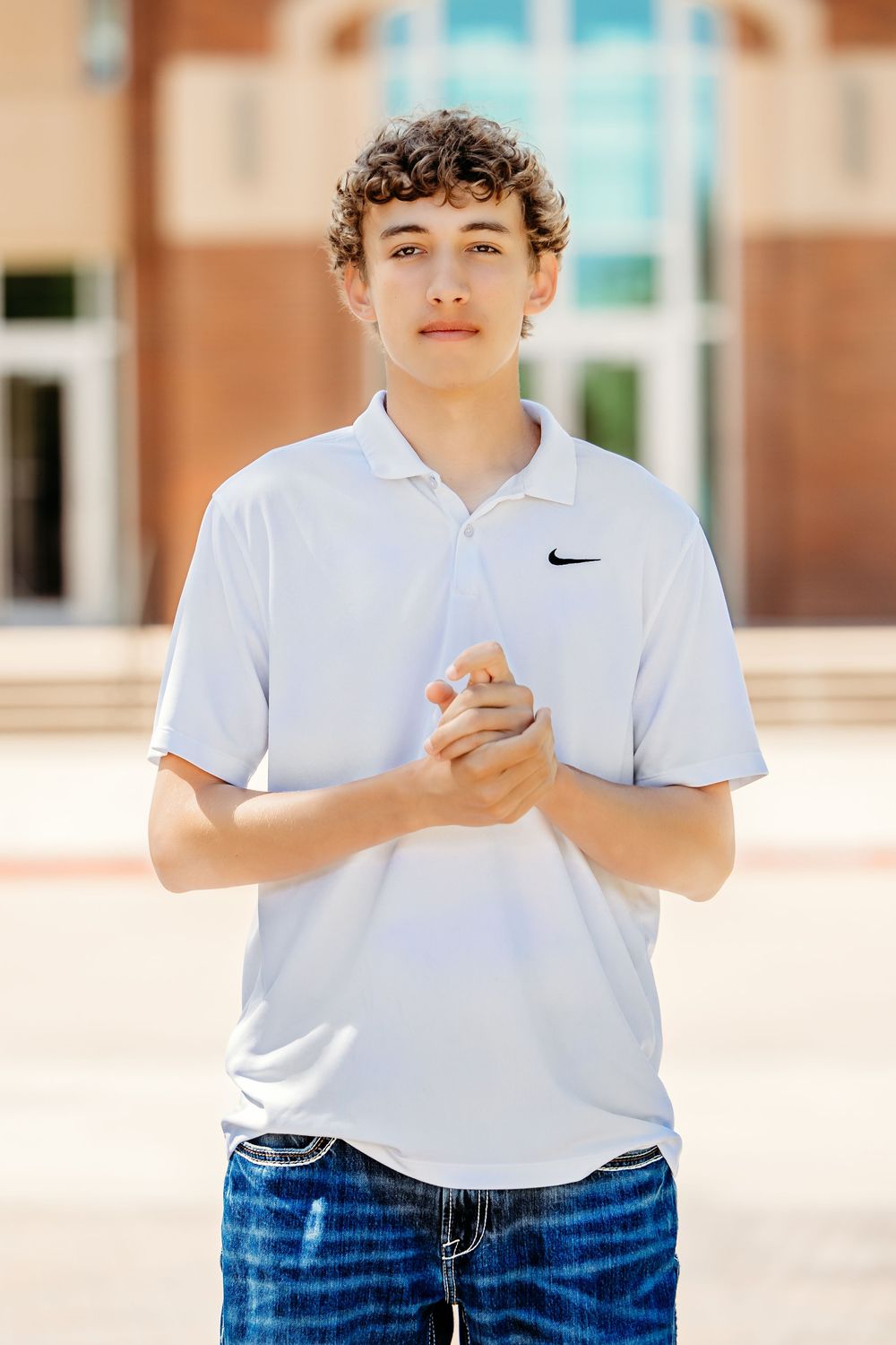 Teen boy in white Nike polo shirt and jeans stands confidently outside a brick building in Keller.