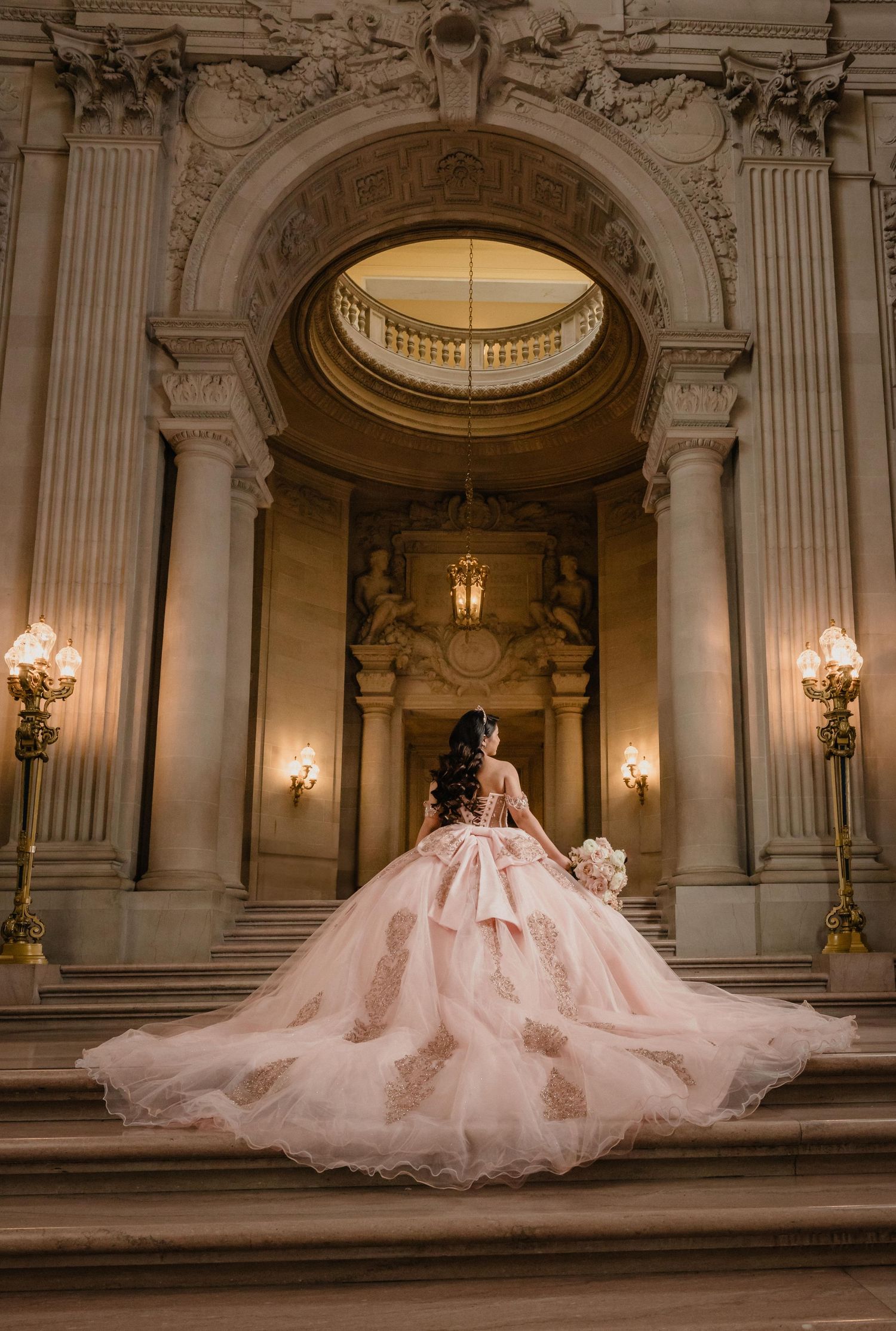 Bride in blush pink ballgown with gold lace stands on grand staircase at San Francisco City Hall.