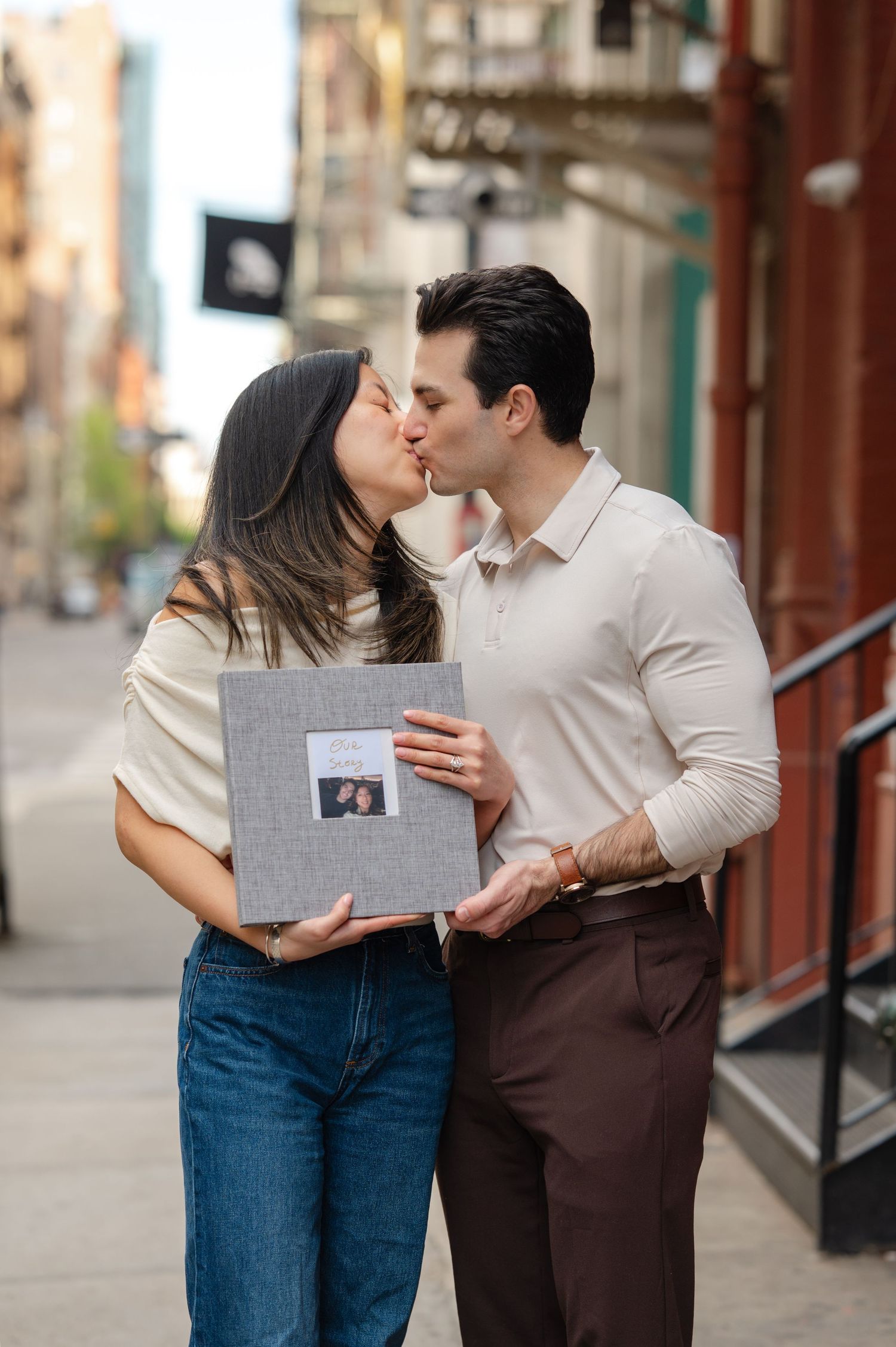 Couple kissing on a city sidewalk while holding a gray photo album with a portrait on the cover with the title "Story of Us" after proposal