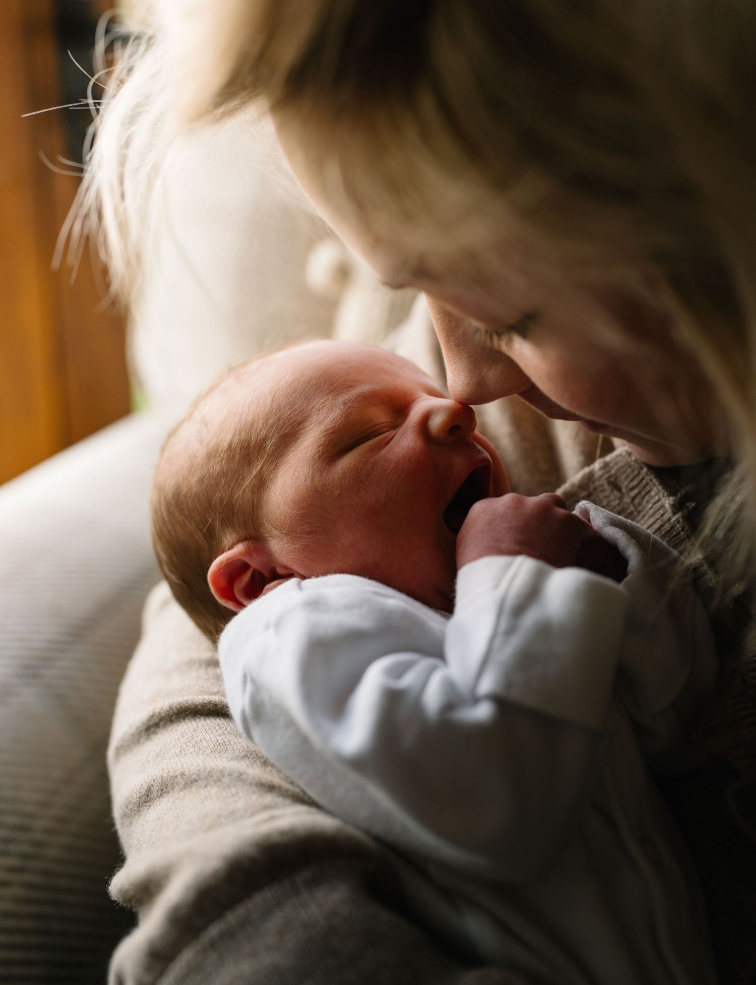 A newborn baby smiles while being cradled in a mother's arms near a bright window.
