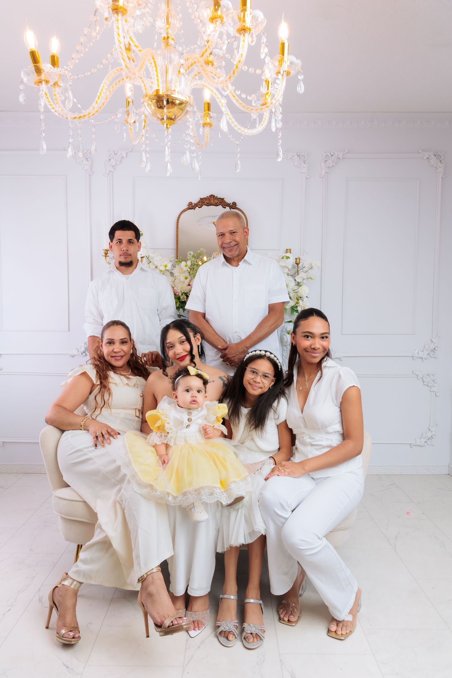 Family portrait in white outfits with baby in yellow dress under crystal chandelier in elegant white room.