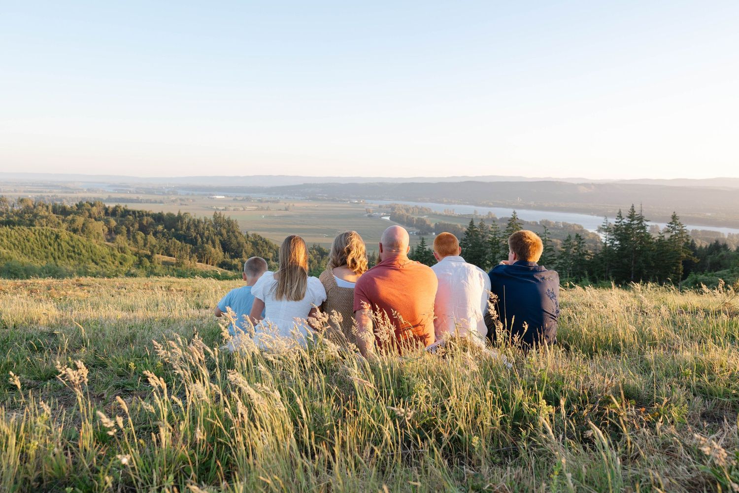 Group of people sitting together on a grassy hilltop overlooking a scenic valley at sunset.