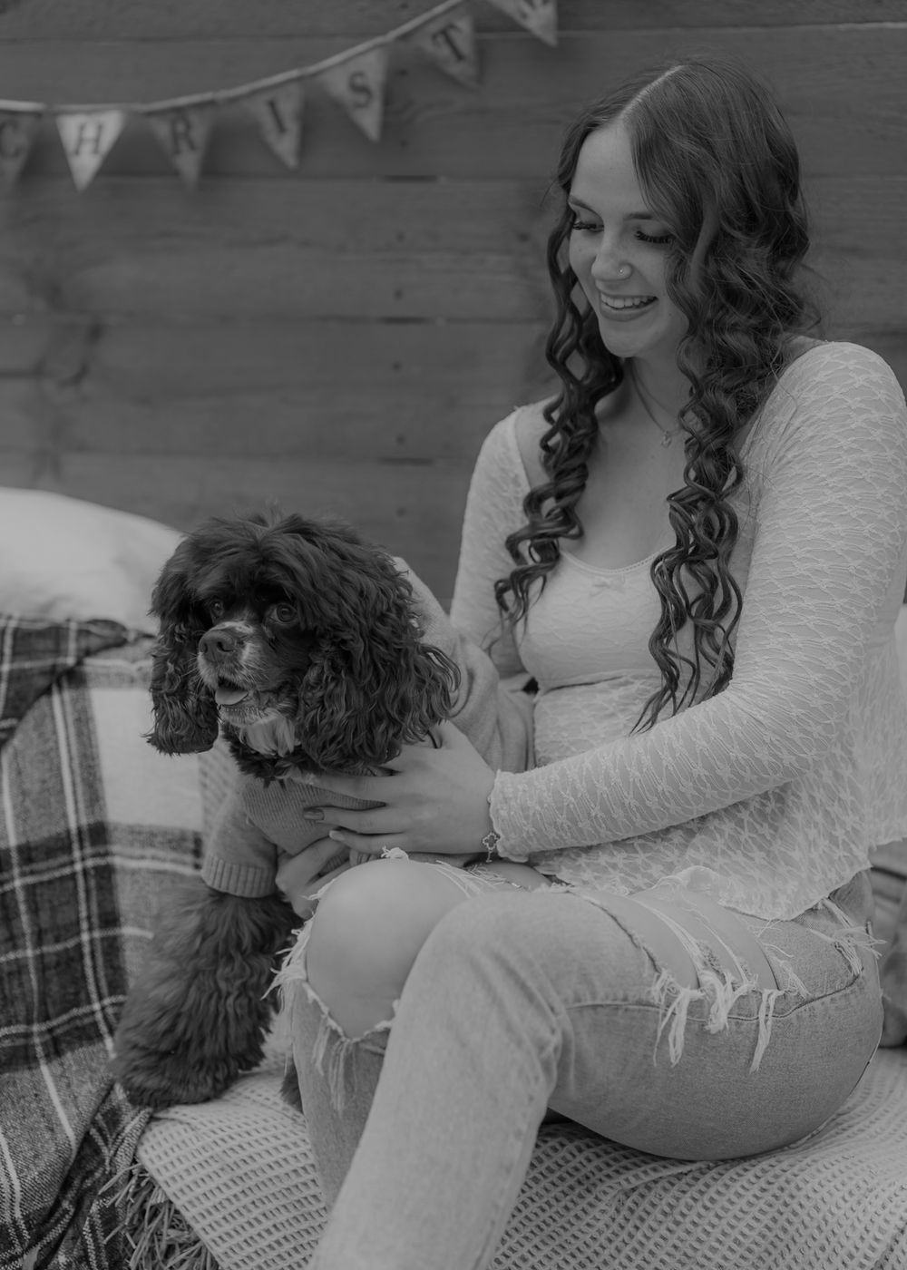A smiling young woman with curly hair sits holding a small dog in a sweater, with a Christmas banner in the background.