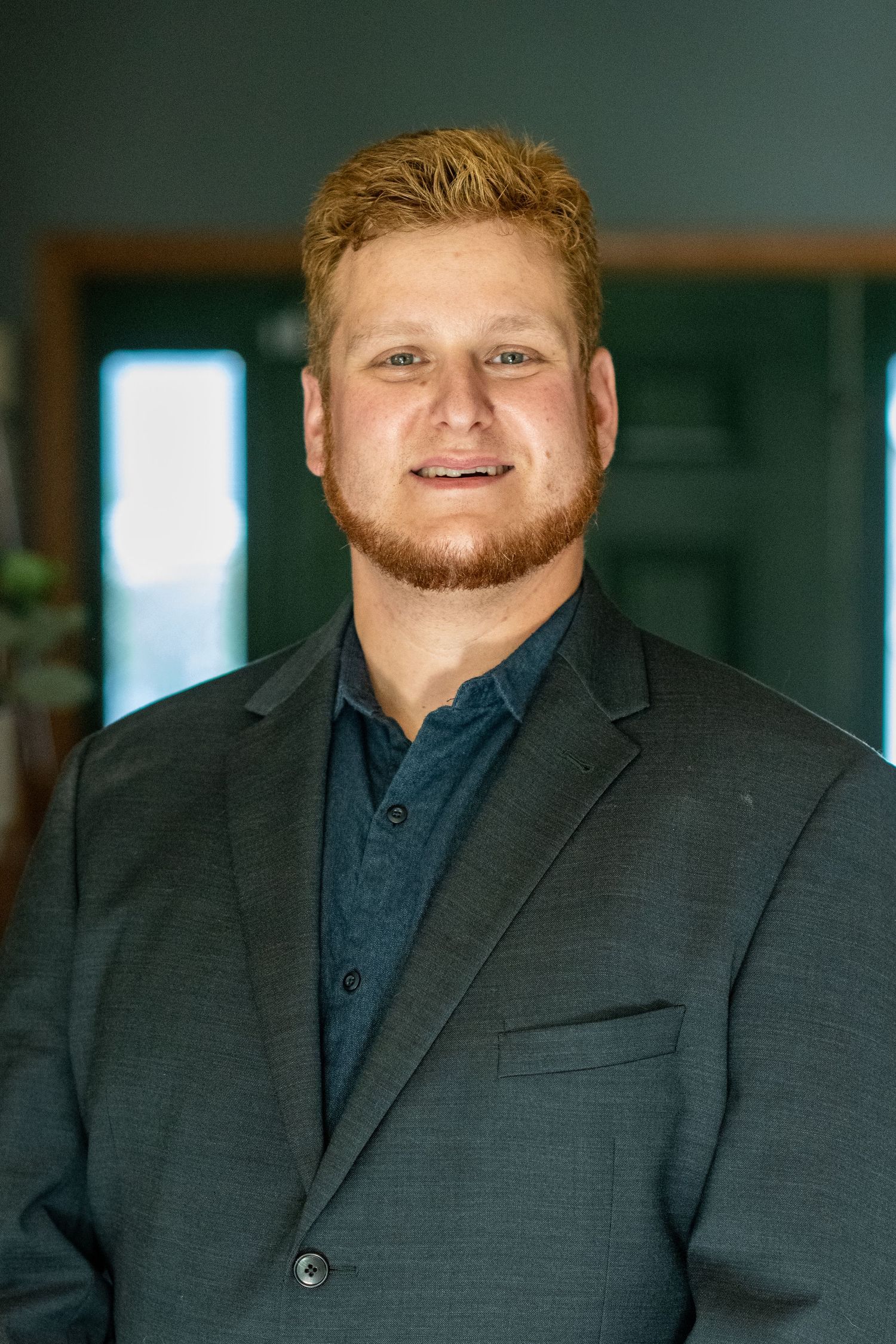 Professional headshot of a young man with red hair and beard wearing a black shirt against a dark green background.