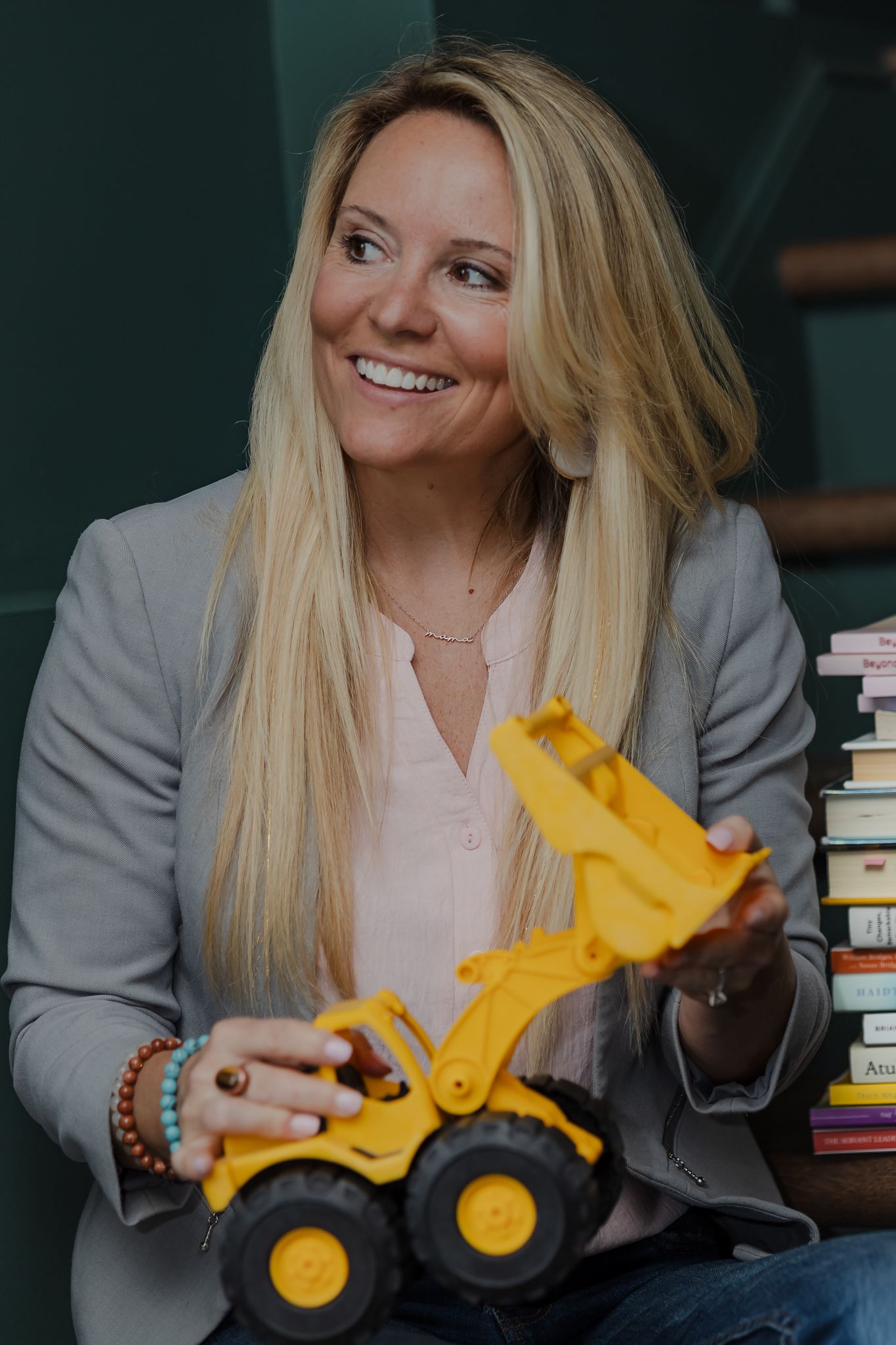 Smiling blonde woman in gray blazer holding yellow toy construction loader near stack of colorful books.