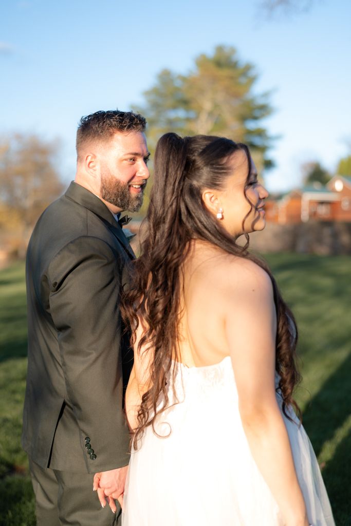A smiling bride and groom in wedding attire hold hands outdoors on a sunny day with green grass and trees behind them.