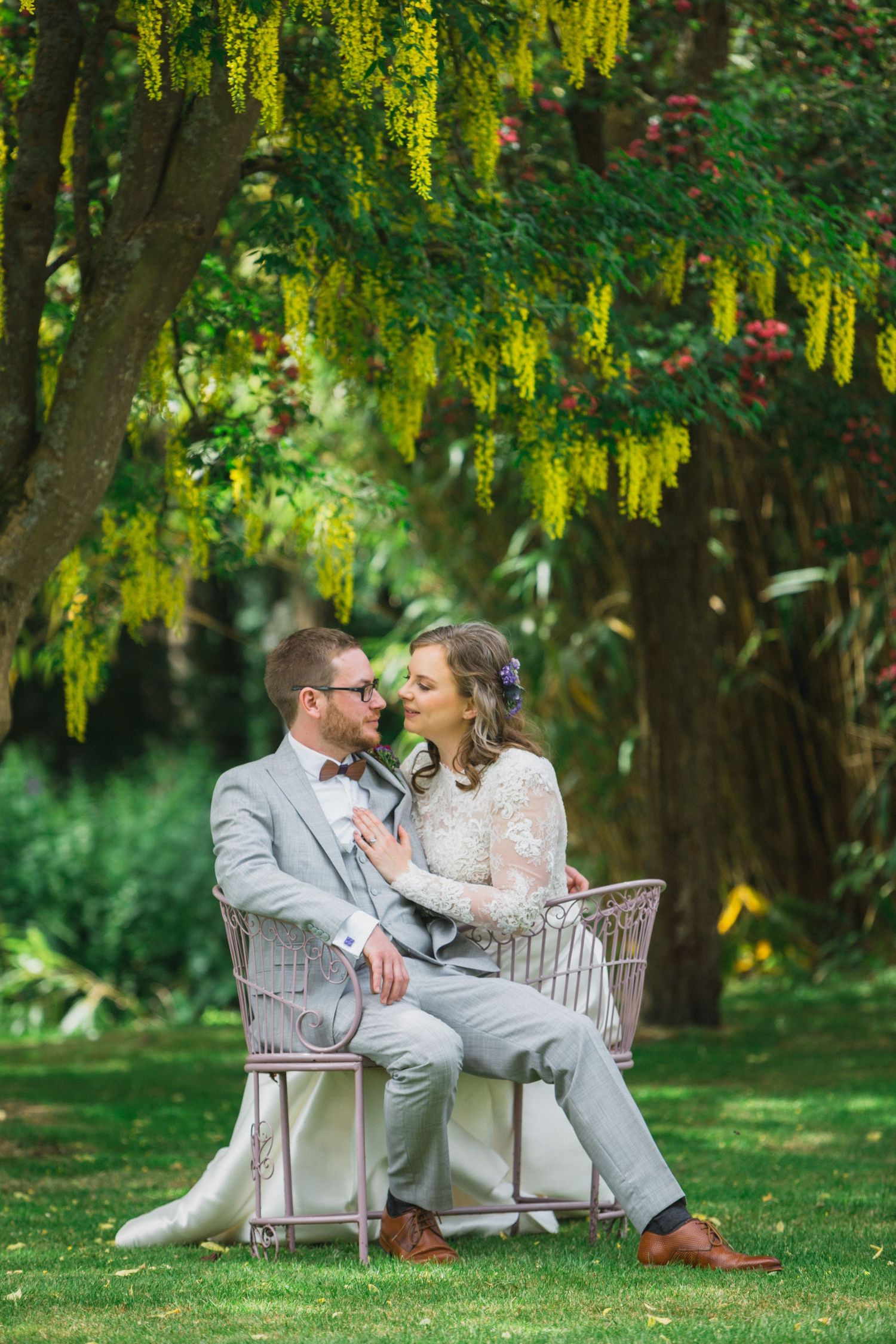 Bride and groom sit together on a vintage chair in a lush garden, gazing at each other under yellow flowering trees.