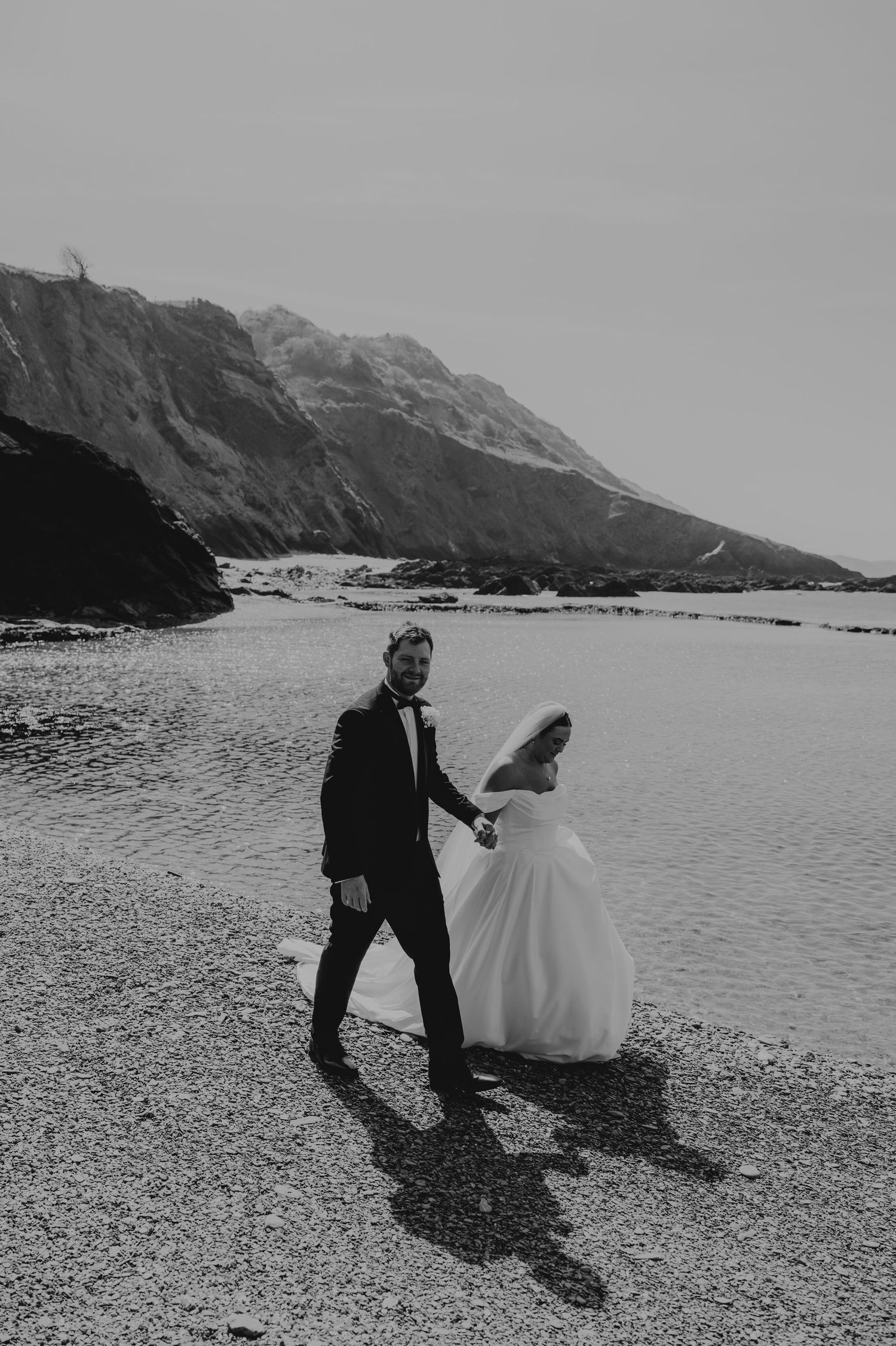 Black and white photo of a bride and groom walking hand in hand along a rocky beach at Tunnels Beaches wedding venue