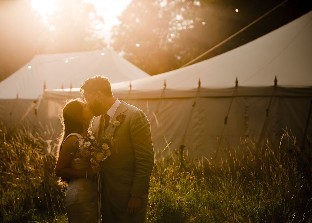 Couple sharing a romantic kiss at golden hour in front of a white marquee tent surrounded by lush greenery.