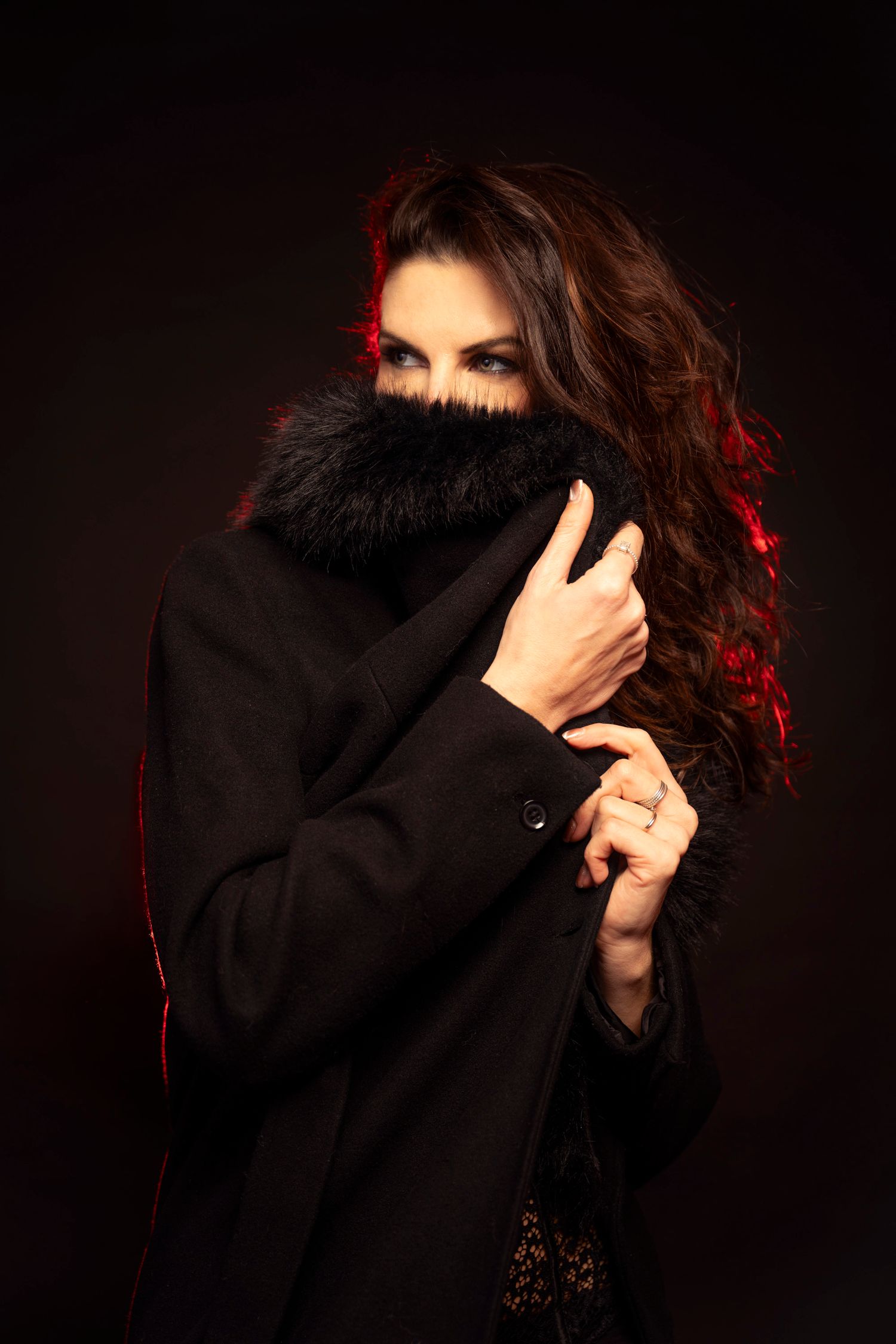Mysterious woman with dark curly hair covering her face with a black fur-collared coat against dark background.